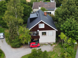 Fågelvy över ett stor hus i tegel och trä. Hustaket lutar och är gjort av svarta takpannor. Intill huset står en röd bil parkerad.