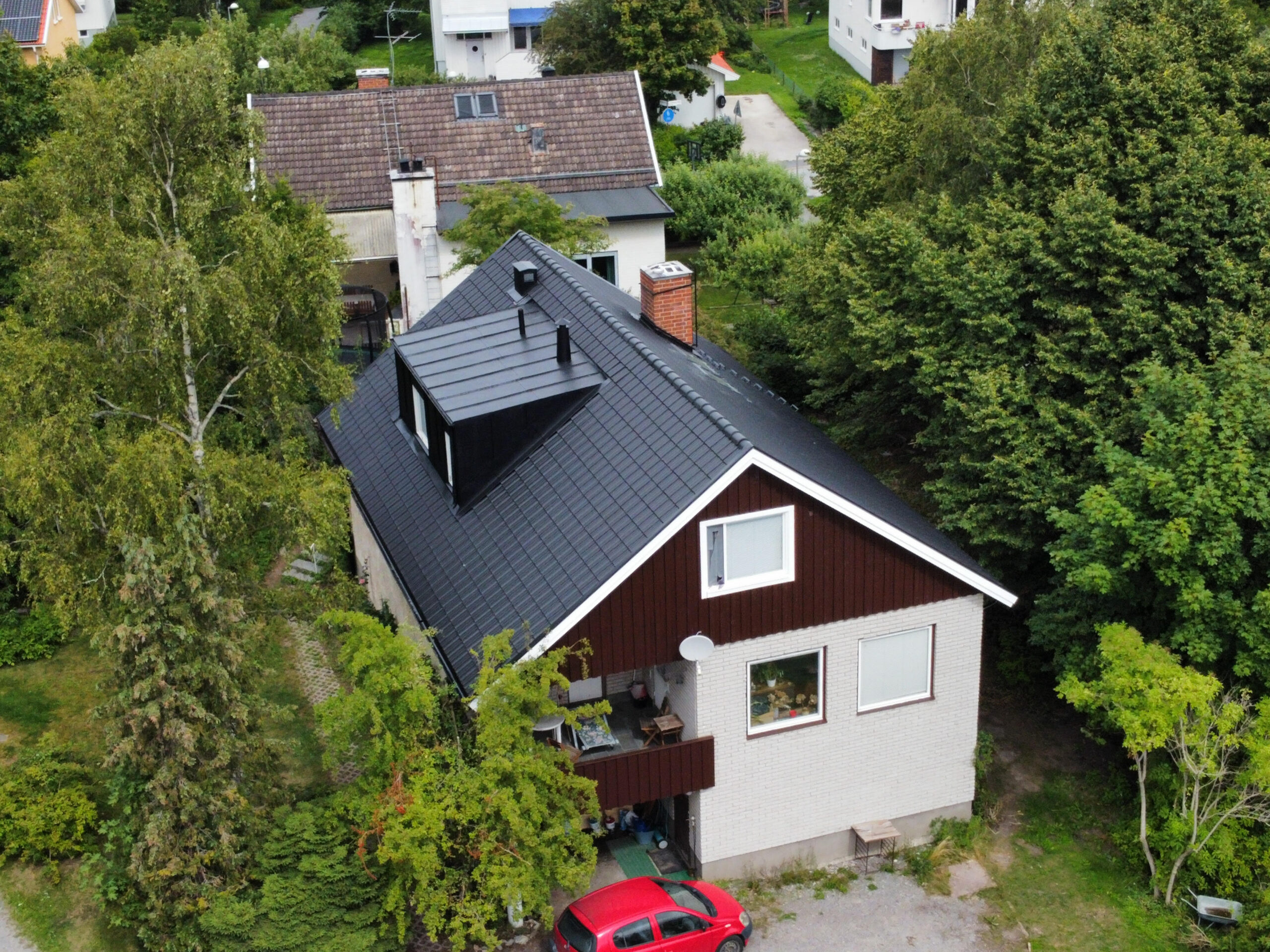 Fågelvy över ett stor hus i tegel och trä. Hustaket lutar och är gjort av svarta takpannor. Fågelvy över ett stor hus i tegel och trä. Hustaket lutar och är gjort av svarta takpannor. Intill huset står en röd bil parkerad.