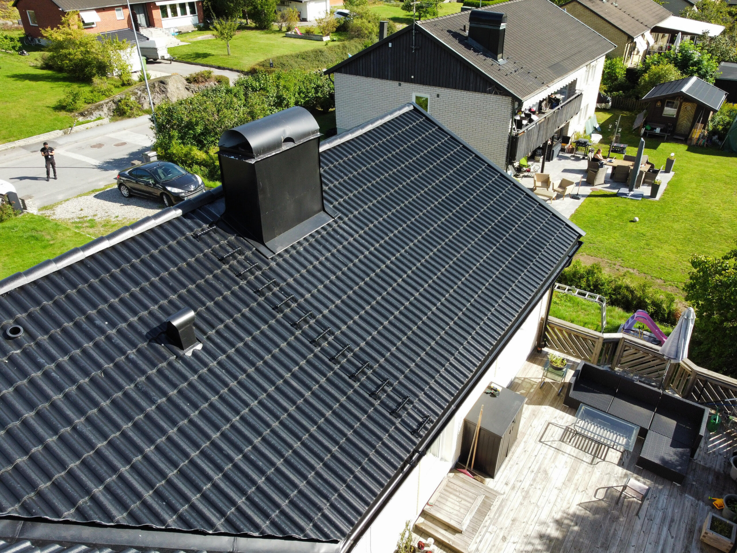 Överblick över hustomt. Huset har ett svart tak av betongpannor. I bakgrunden syns närliggande grannhus.