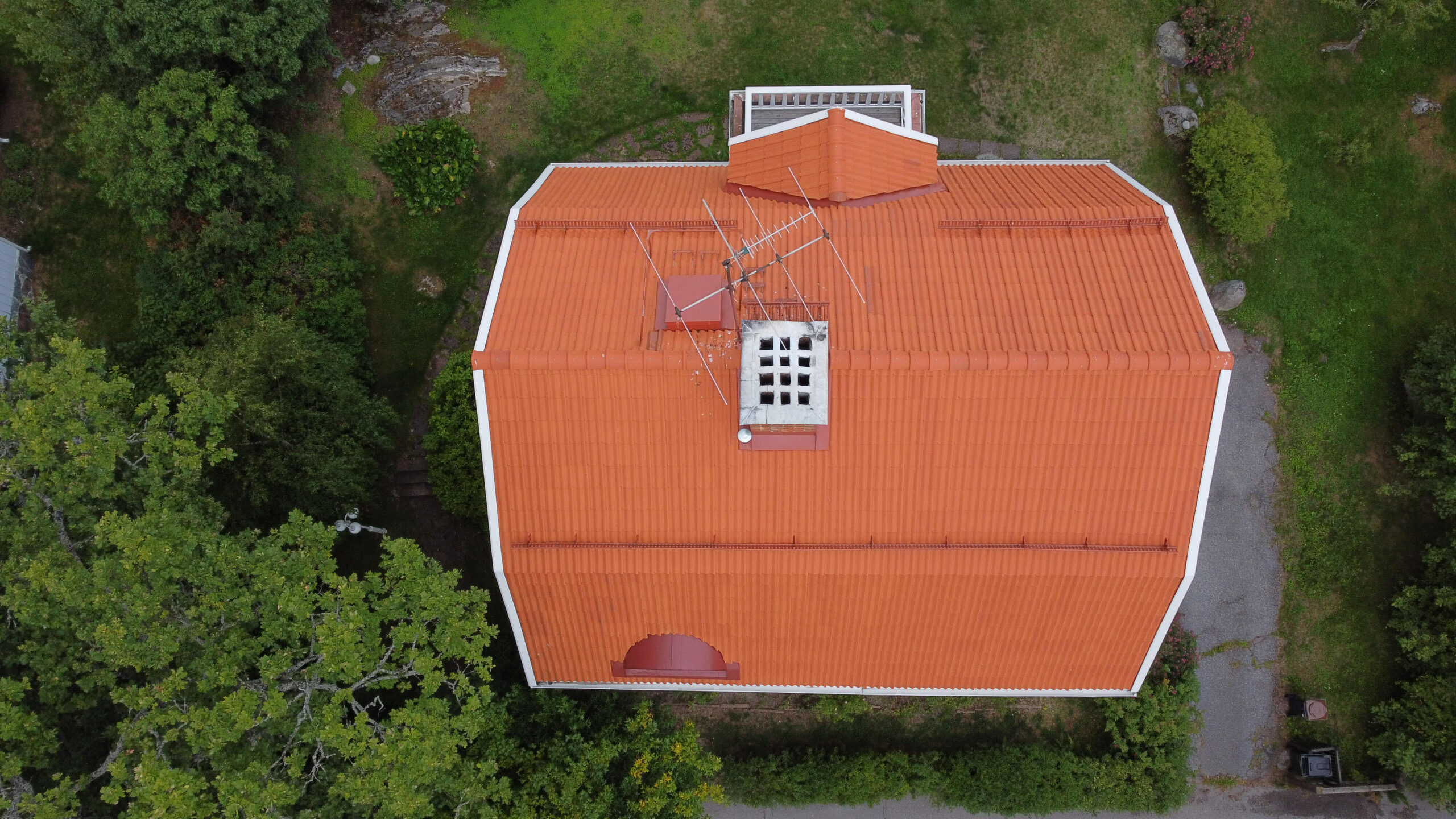 Fågelvy ovanifrån en stor villa med ett lutande tak och orangea tegelpannor. Taket har en skorsten, snörasskydd samt ett vindsfönster.