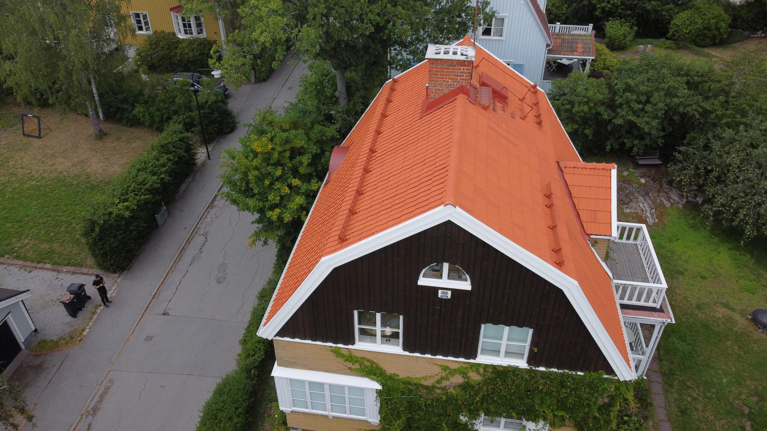 Ett stort villahus i trä och tegel, med sluttande tak och orangea takpannor. Taket har en skorsten, snörasskydd samt ett vindsfönster.