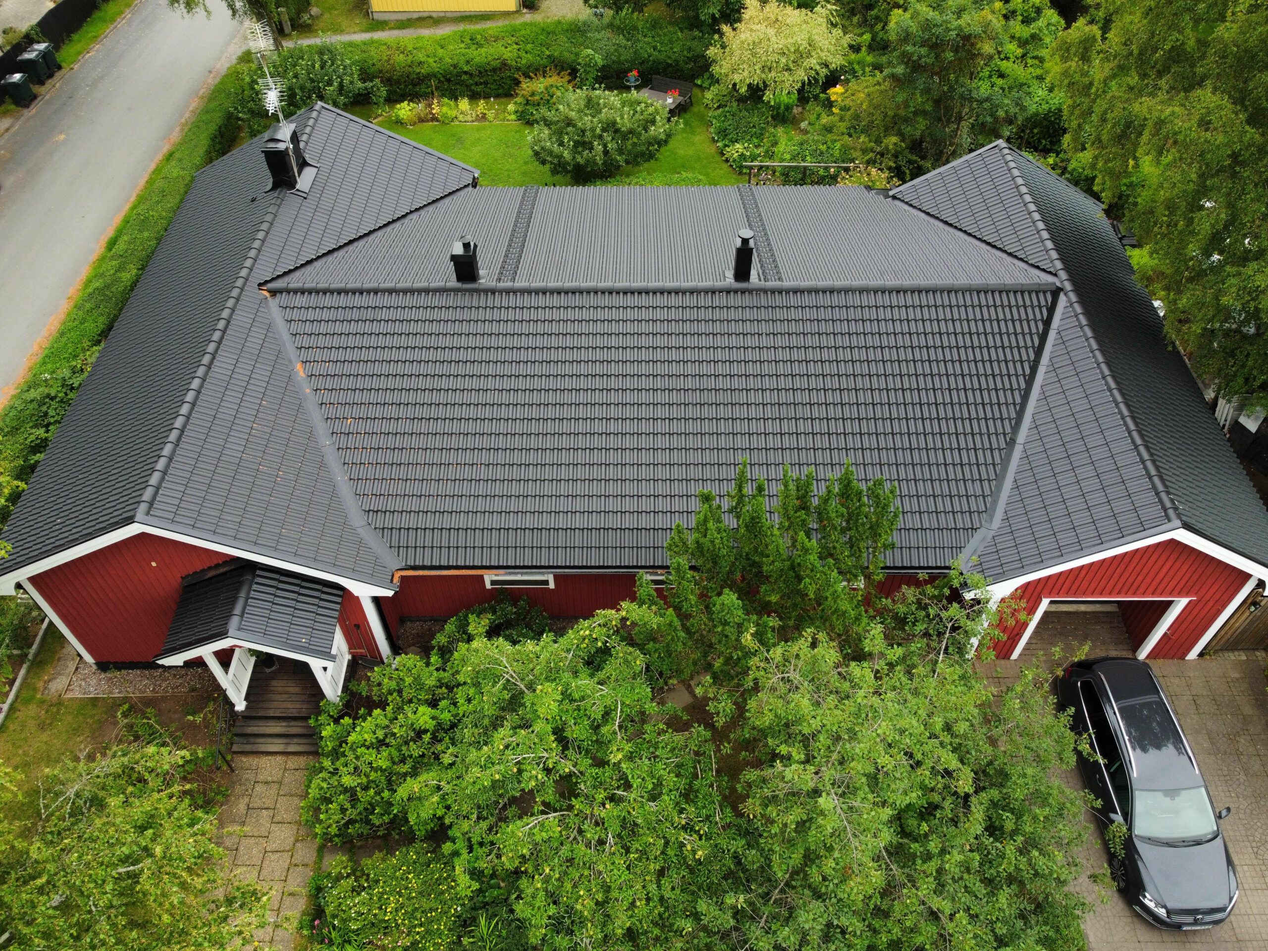 Fågelvy över ett stort rött villa hus, med sluttande tak av svarta betongpannor. På huset finns en skorsten samt två takstegar. Huset är omringat av träd och gräsmatta.