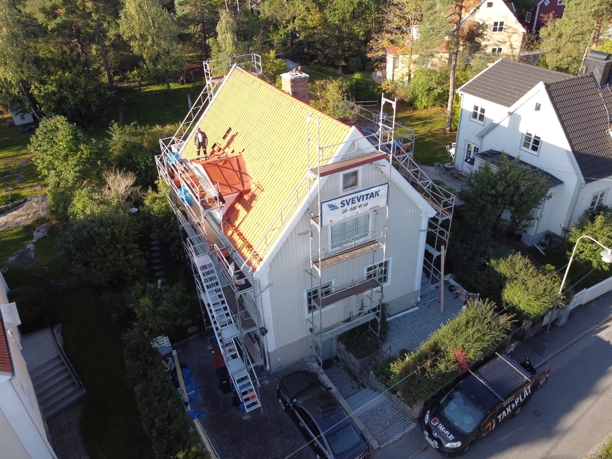 Fågelvy över hustak under takomläggning. Runt huset står flera byggställningar. I bakgrunden syns närliggande grannhus.