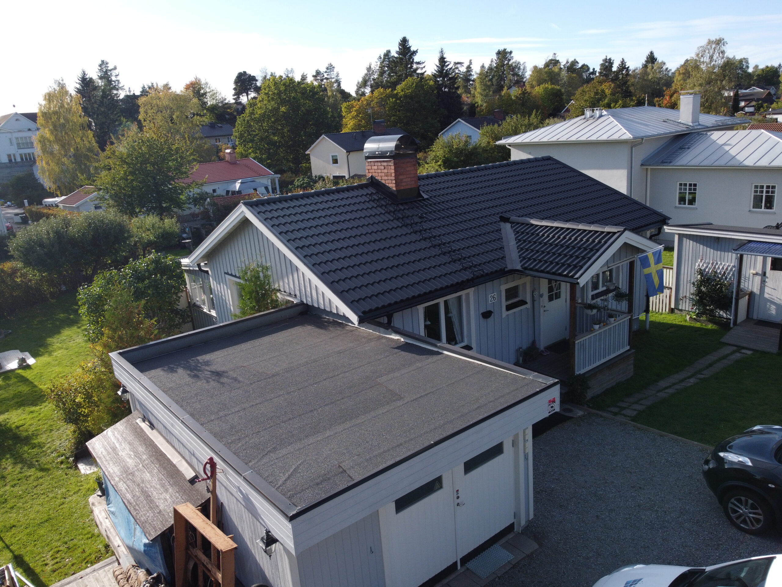 Fågelvy över hustak efter takomläggning. Taket är gjort av svarta betongpannor, och har en skorsten i tegel. Tillhörande garage har platt papptak. I bakgrunden syns närliggande grannhus.