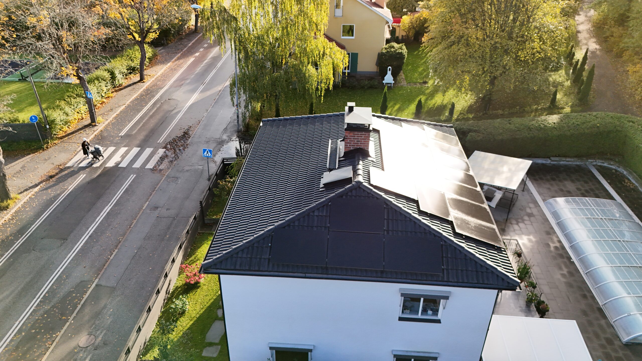 Fågelvy över hustak efter takomläggning med betongpannor och solceller. Taket har en skorsten i tegel samt snörasskydd. Nedanför taket syns hustomten och närliggande grannhus.