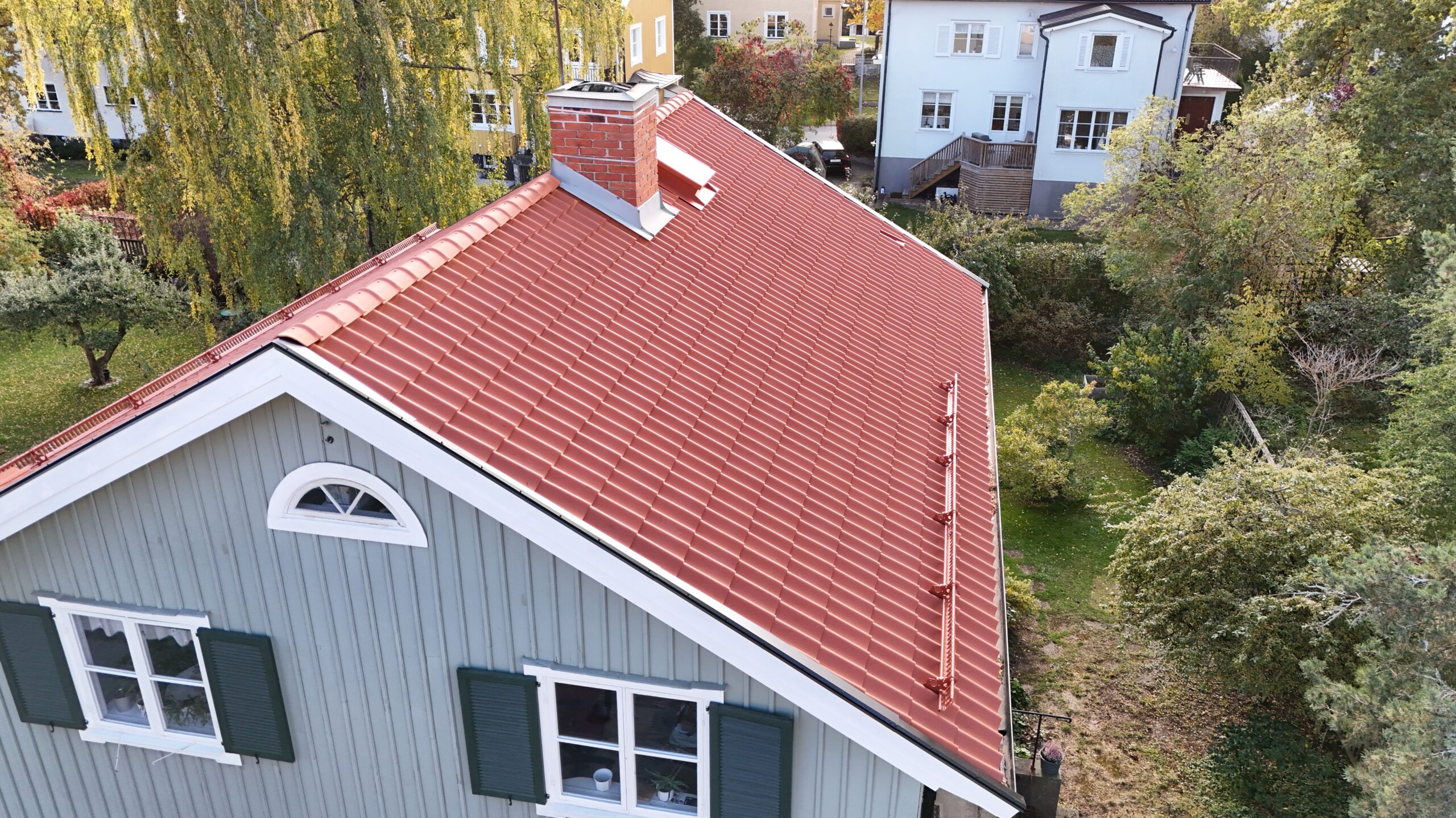 Fågelvy över en grön villa med sluttande tak av tegelpannor. På taket finns en skorsten i tegel samt snörasskydd. I bakgrunden syns närliggande grannhus.