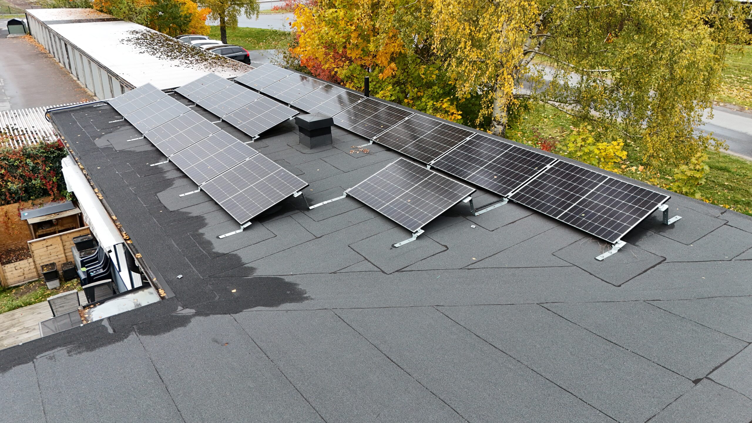 Närbild ovanpå ett radhus med solceller på papptaket. I bakgrunden syns områdets garage samt några träd.