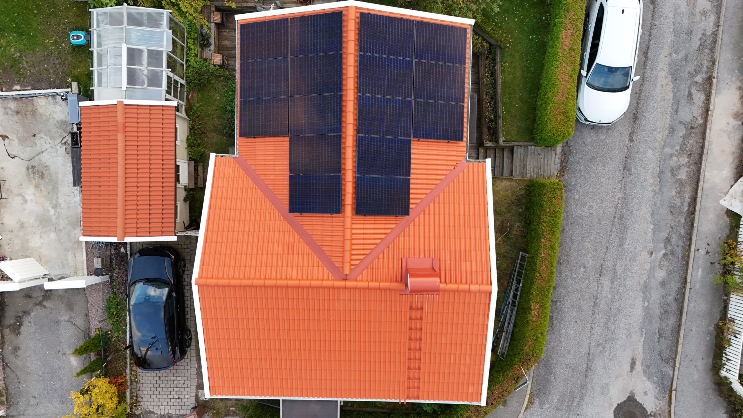 Fågelvy över hustak efter takomläggning. Hustaket är av tegelpannor, och har solceller, en takstege samt en skorsten i plåt. Runt huset syns delar av hustomten, samt två parkerade bilar.