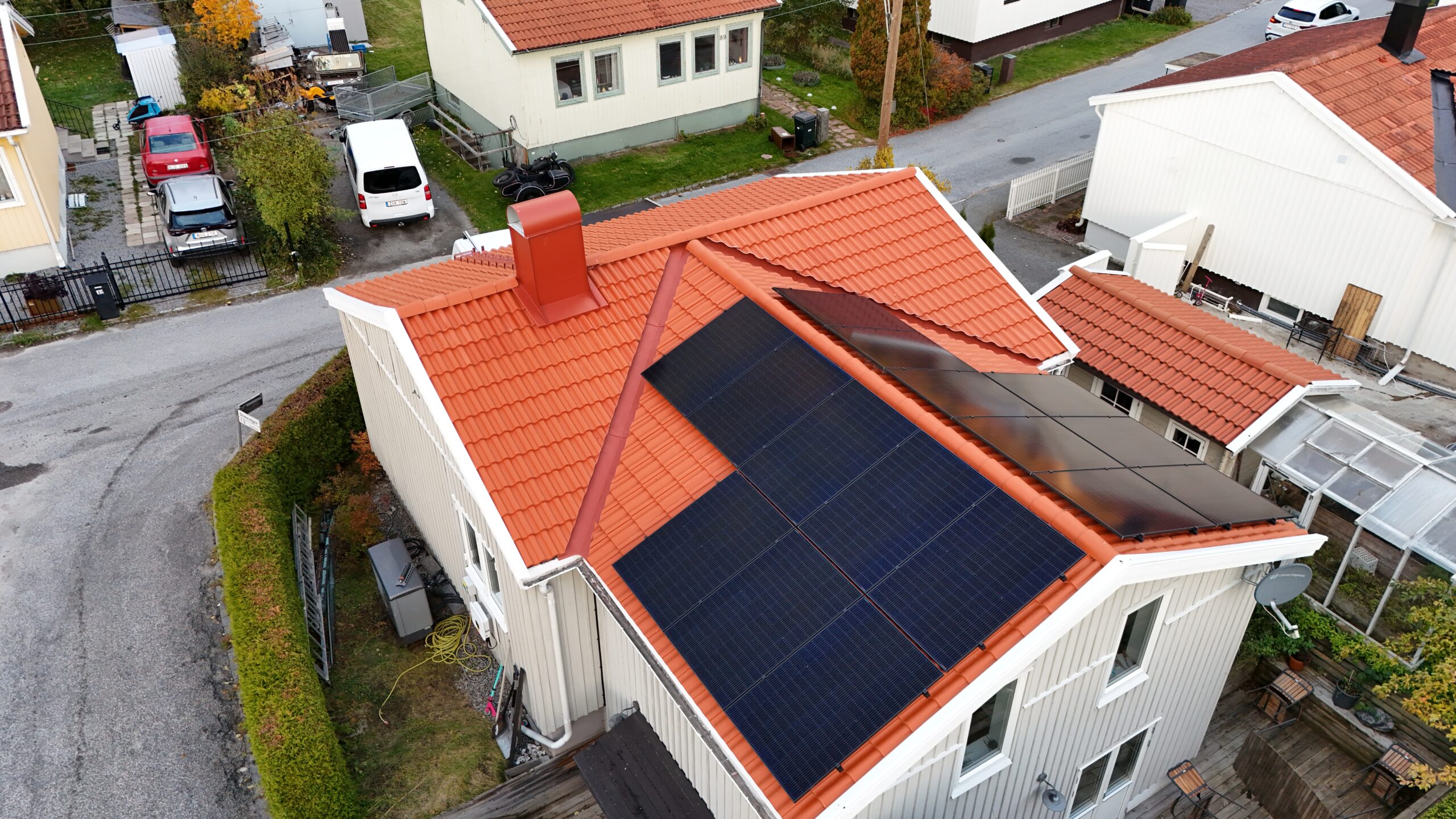 Fågelvy över hustak efter takomläggning. Hustaket är av tegelpannor, och har solceller, en takstege samt en skorsten i plåt. Runt huset syns närliggande grannhus.