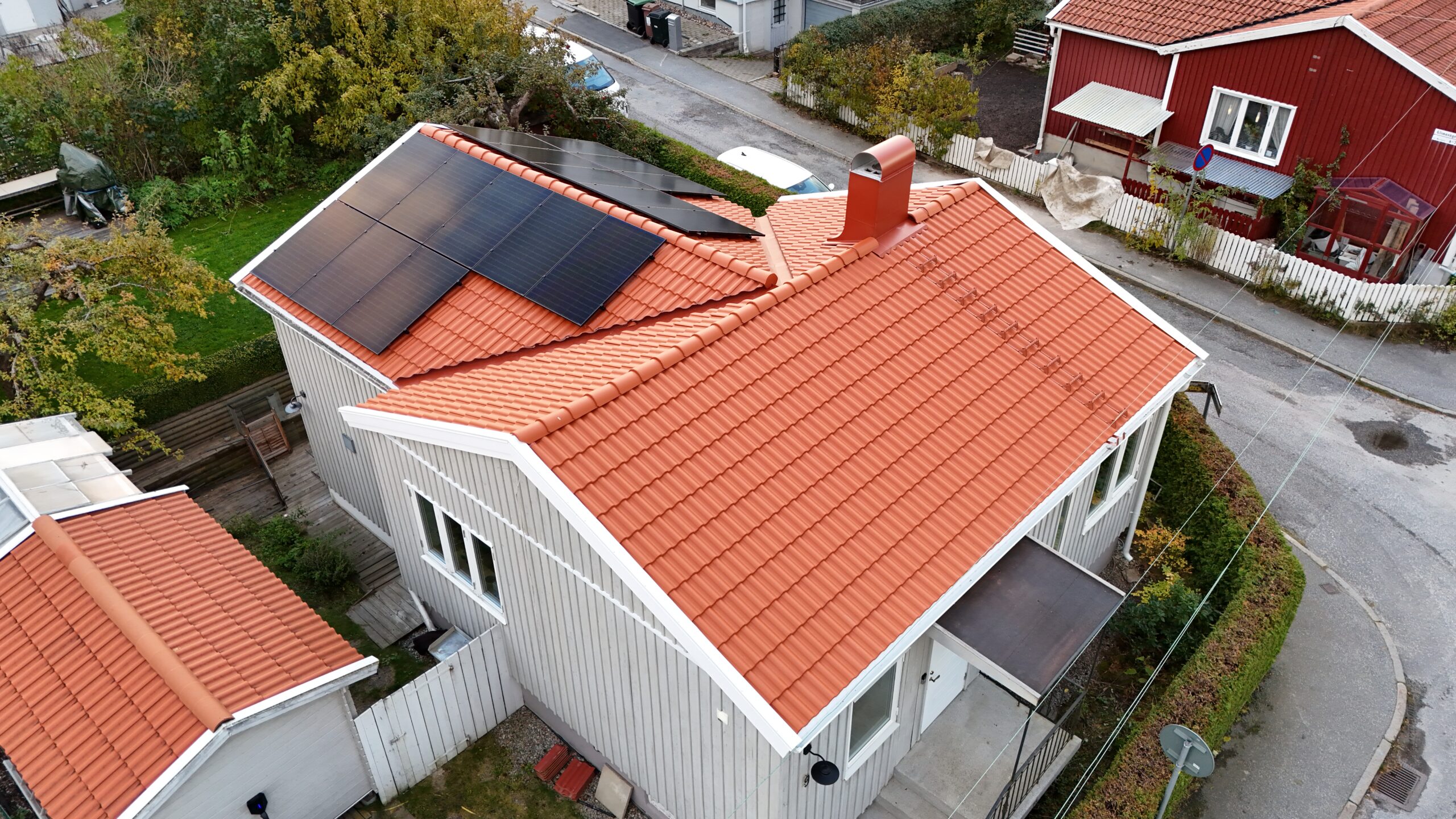 Fågelvy över hustak efter takomläggning. Hustaket är av tegelpannor, och har solceller, en takstege samt en skorsten i plåt. Runt huset syns närliggande grannhus.