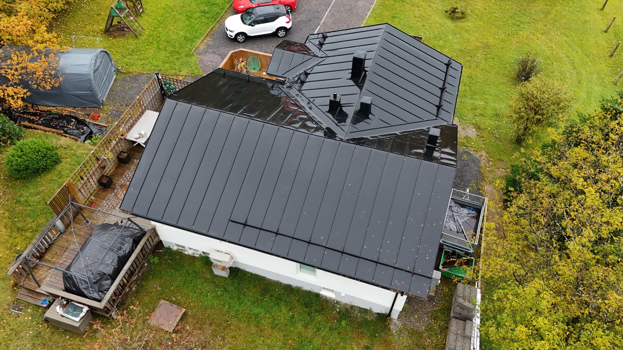 Fågelvy över ett vitt hus med svart, bandtäckt plåttak. Huset står på en tomt med gräsmatta och bilar.