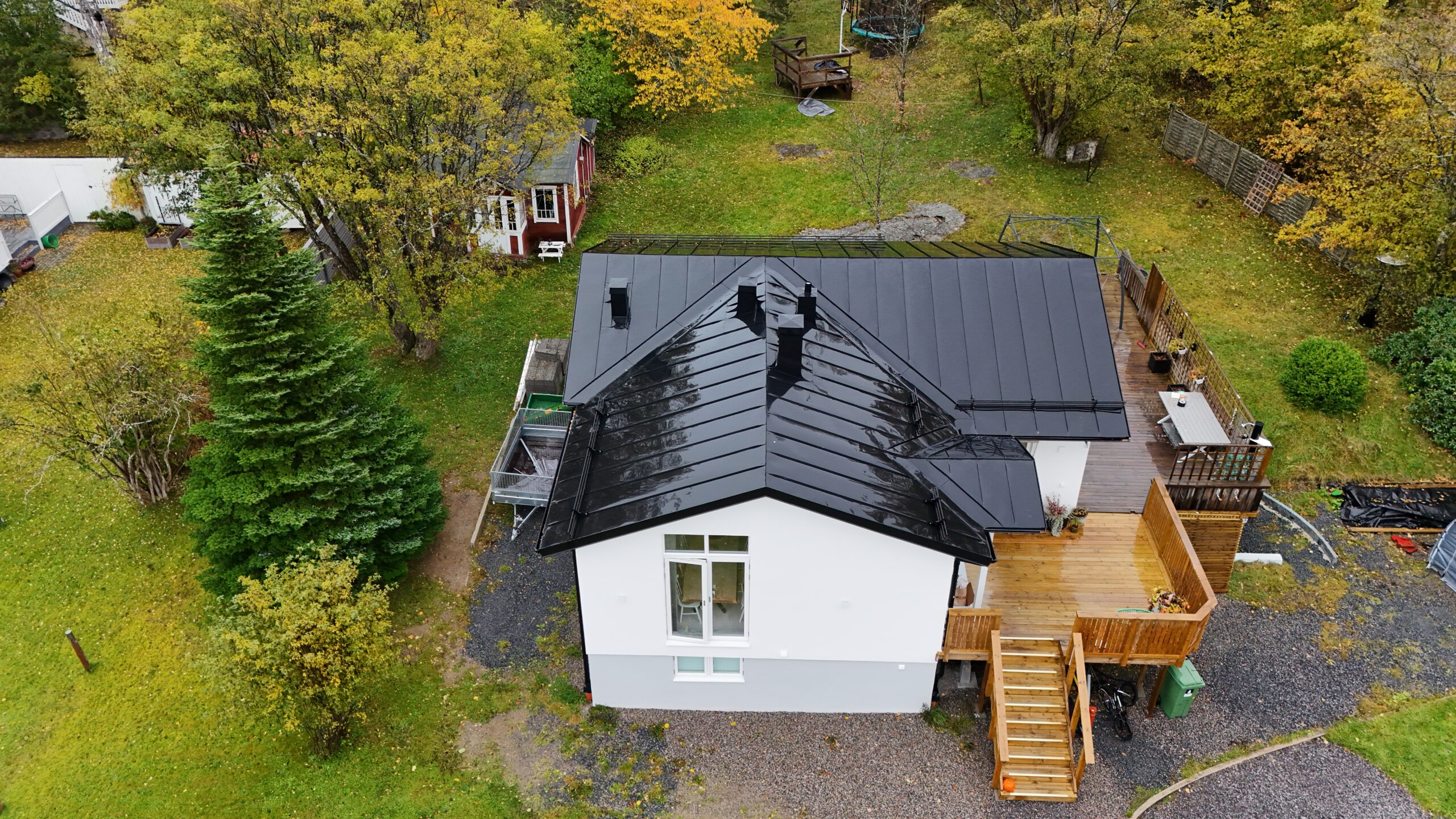 Fågelvy över ett vitt hus med svart, bandtäckt plåttak. I bakgrunden syns hustomten.