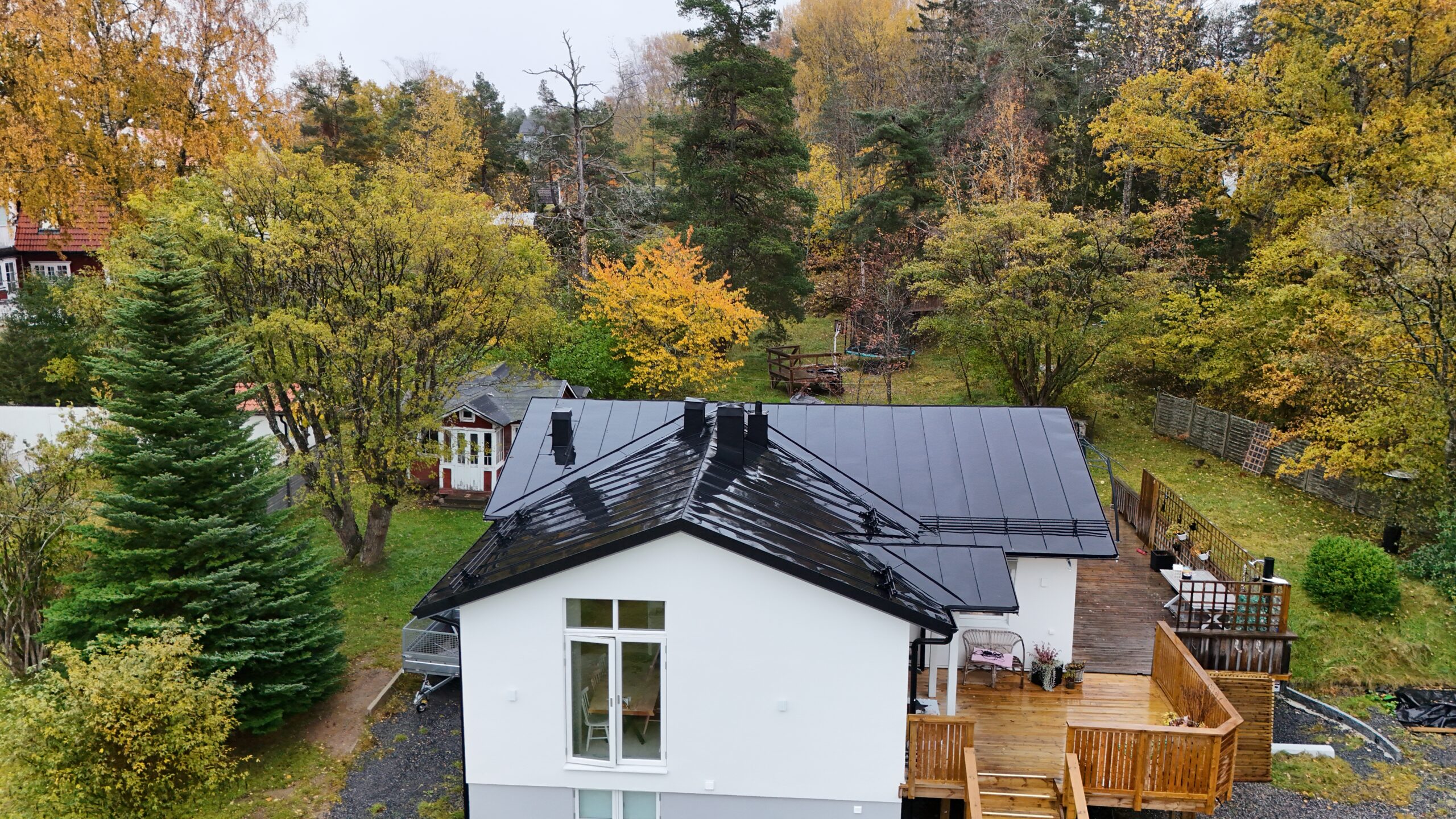 Vy över ett vitt betonghus med sluttande bandtäckt plåttak. Huset är omringat av träd på tomten.