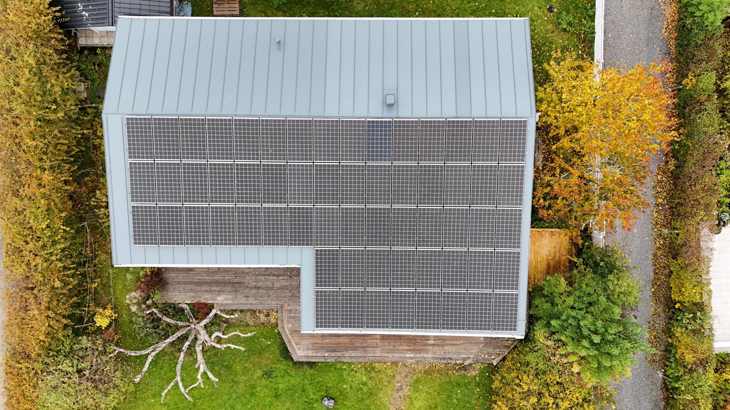 Fågelvy över taket på ett vitt enplanshus. Hustaket är gjort av bandtäckt plåt, och täcks med en stor panel av solceller. Nedanför huset syns en altan och tillhörande hustomt.