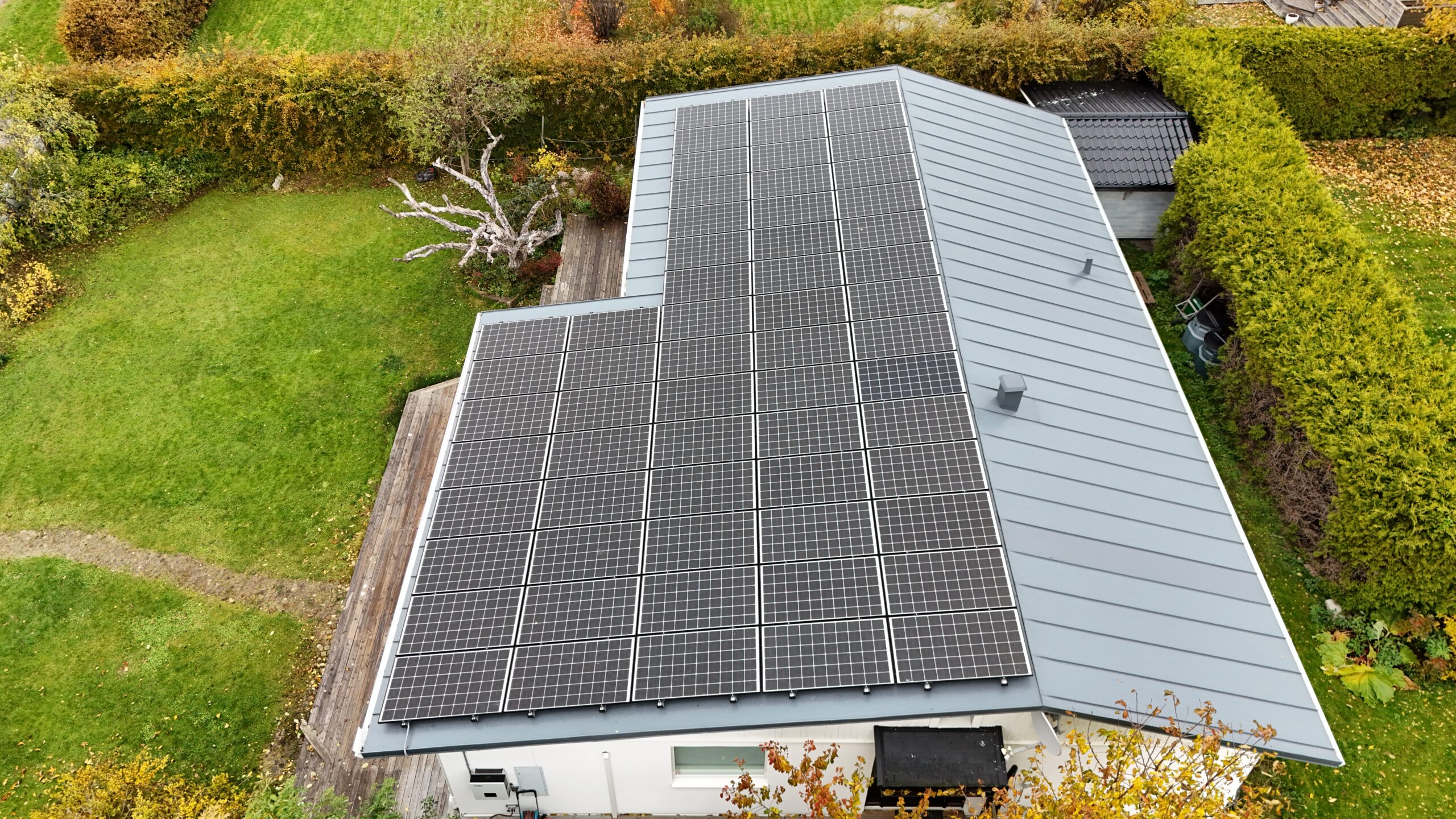 Fågelvy över taket på ett vitt enplanshus. Hustaket är gjort av bandtäckt plåt, och täcks med en stor panel av solceller. Nedanför huset syns en altan och tillhörande hustomt.