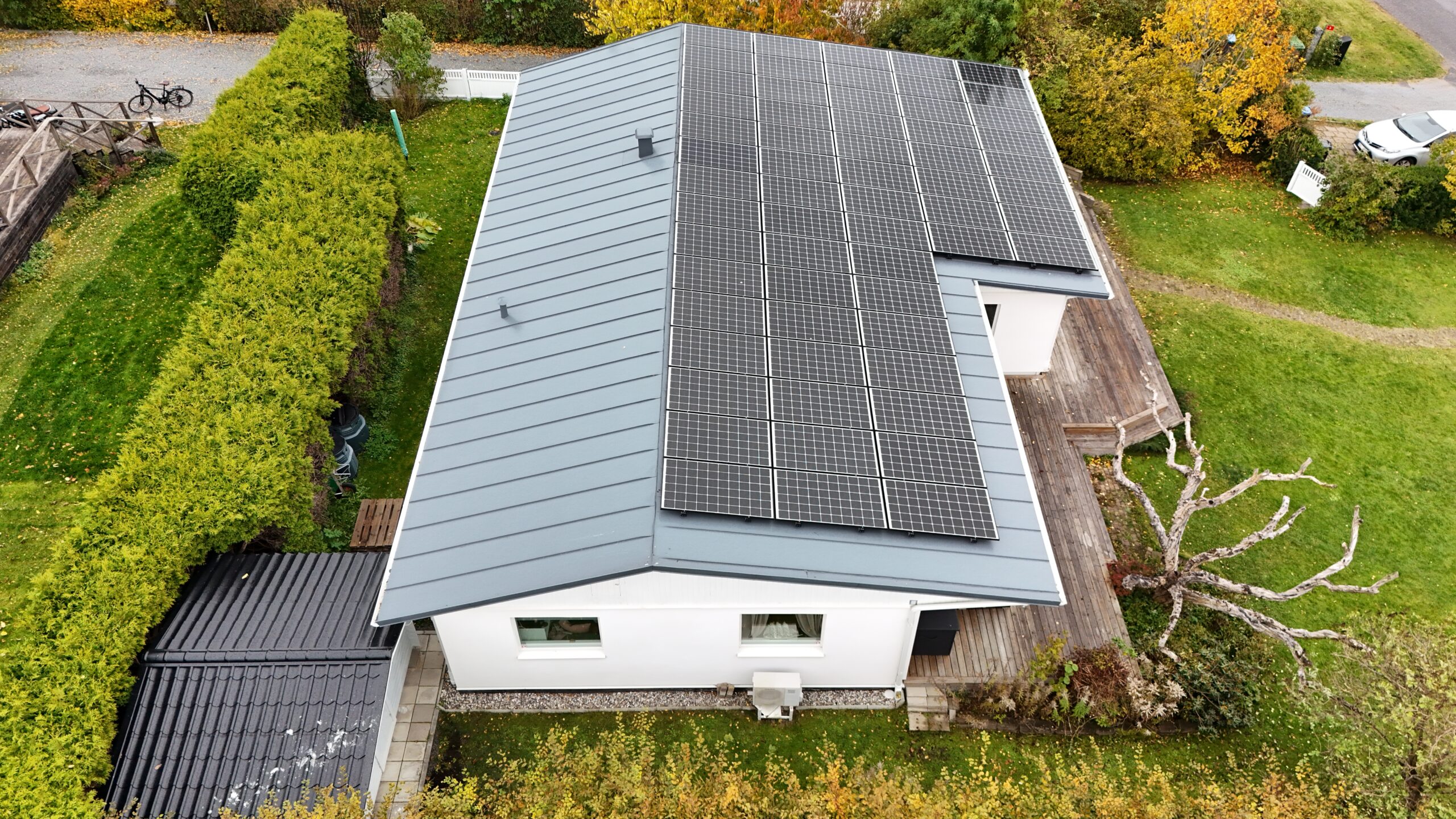 Fågelvy över taket på ett vitt enplanshus. Hustaket är gjort av bandtäckt plåt, och täcks med en stor panel av solceller. Nedanför huset syns en altan och tillhörande hustomt.