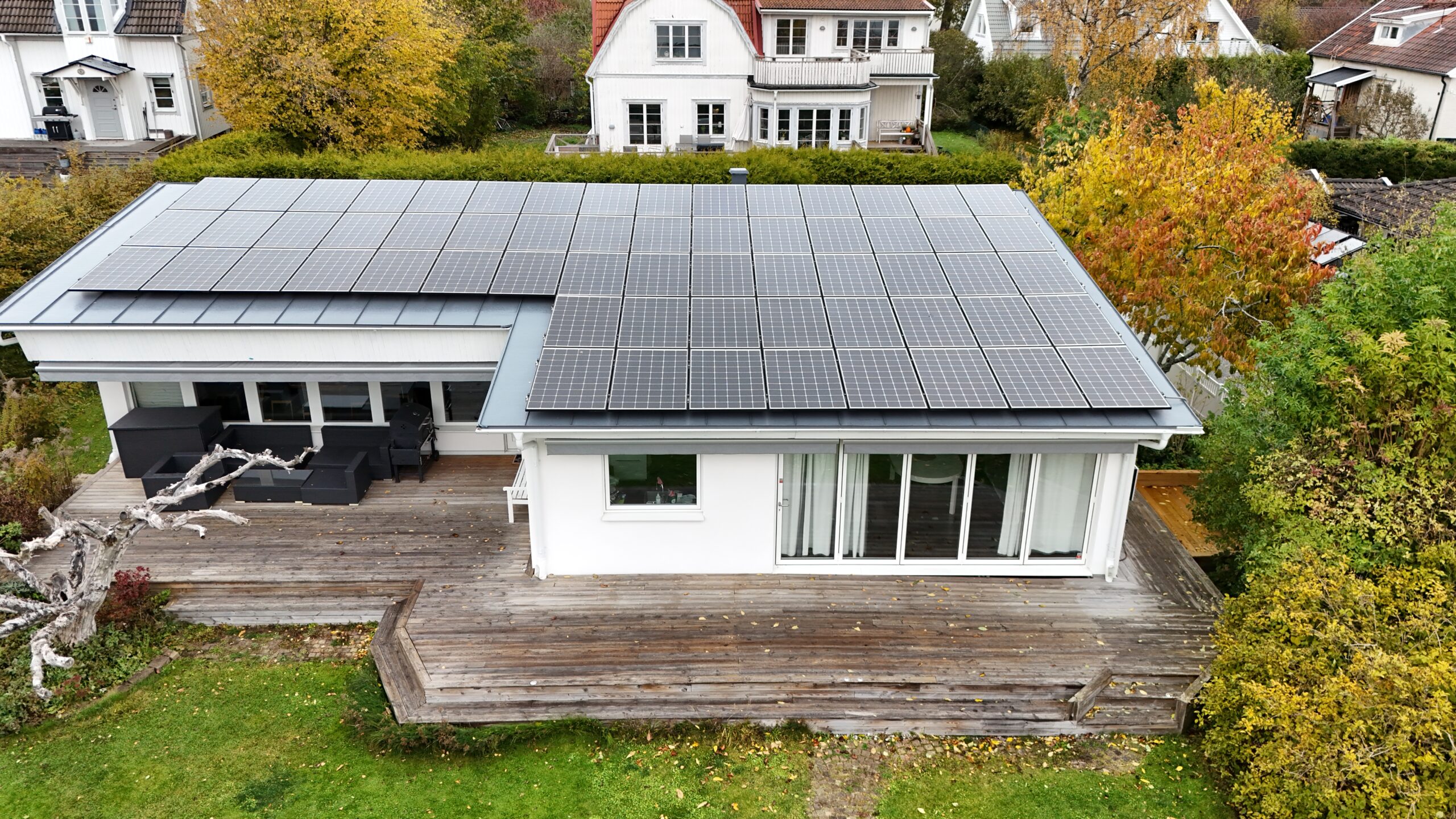 Fågelvy över långsidan på ett vitt enplanshus med omlagt tak. Hustaket är gjort av bandtäckt plåt, och täcks med en stor panel av solceller. Nedanför huset syns en altan och gräsmatta. I bakgrunden syns närliggande grannhus.