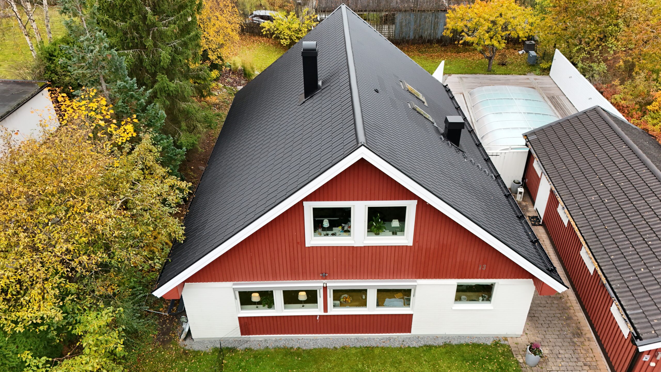 Fågelvy över kortsidan på en vit och röd villa med ett omlagt hustak. Taket är gjort med svarta betongpannor, och har en skorsten, 2 små fönster, snörasskydd samt en takstege.