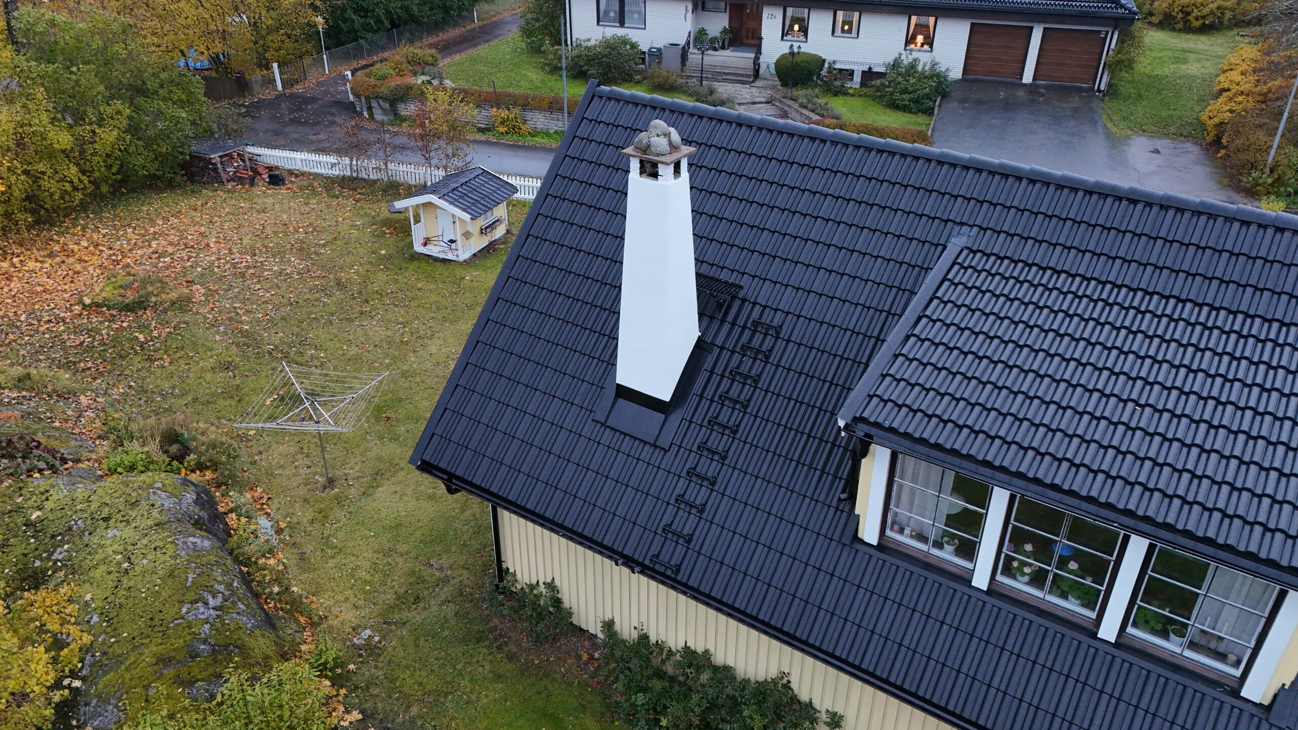 Närbild på ett hustak med omlagda betongpannor. På taket finns en vit skorsten, snörasskydd samt en takstege. Mitt på taket finns en utbuktning med tre fönster. I bakgrunden syns tillhörande hustomt samt ett grannhus.