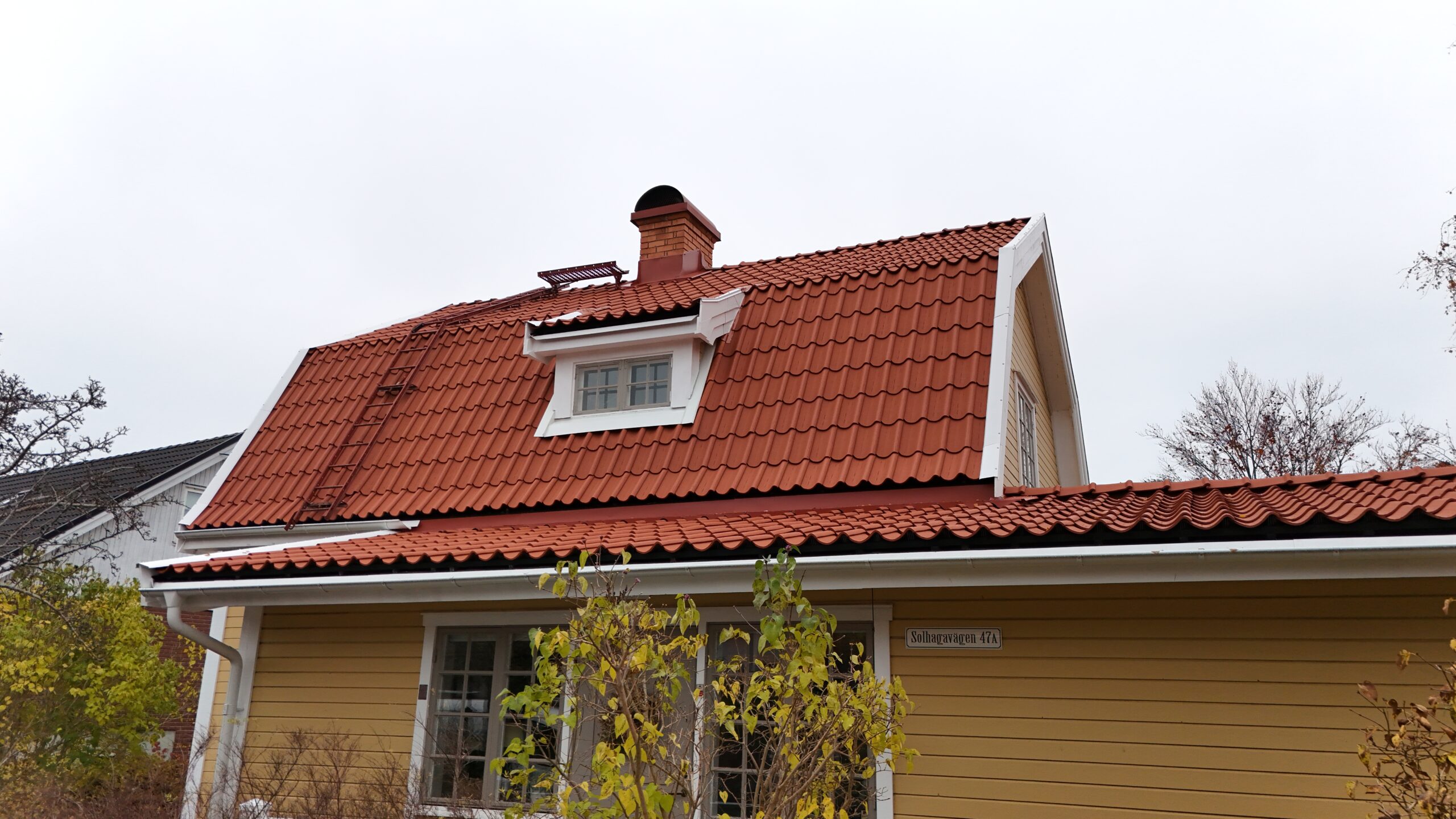 Närbild på långsidan av en gul villa med omlagt tegeltak. På taket finns det en skorsten av tegel, en takstege samt snörasskydd.