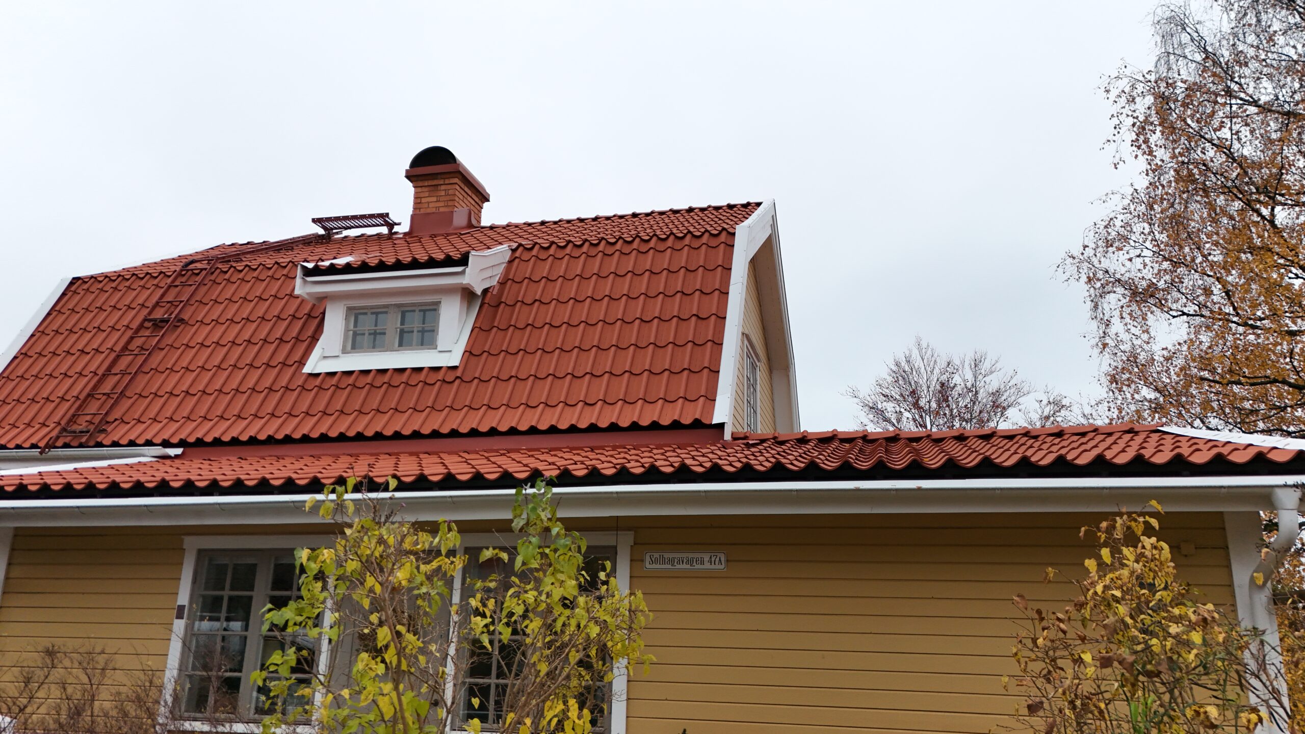 Närbild på långsidan av en gul villa med omlagt tegeltak. På taket finns det en skorsten av tegel, en takstege samt snörasskydd.