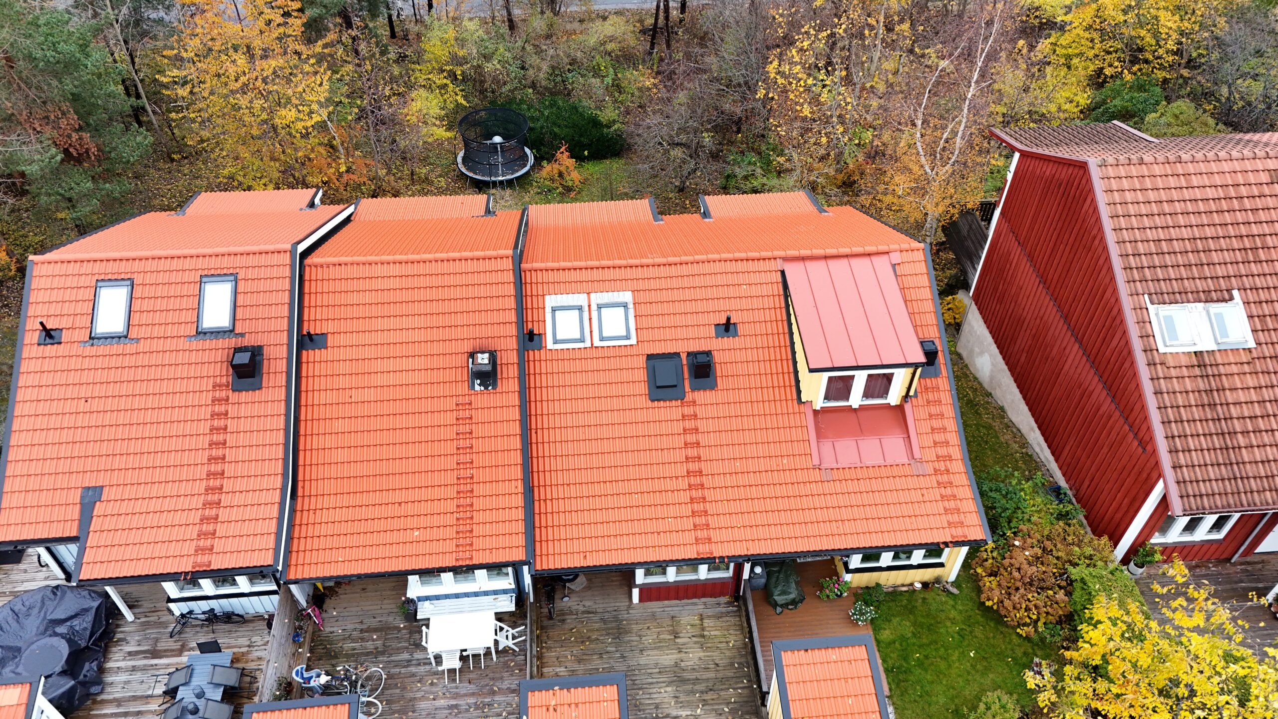 Fågelvy över radhuslänga som genomgått takomläggning. Taket är lätt sluttande, gjort med tegelpannor och detaljer i bandtäckt plåt. På taken finns små fönster, skorstenar och takstegar.