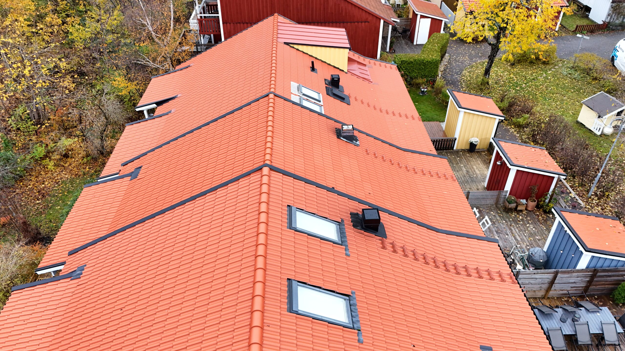 Närbild på radhuslänga som genomgått takomläggning. Taket är lätt sluttande, gjort med tegelpannor och detaljer i bandtäckt plåt. På taken finns små fönster, skorstenar och takstegar.