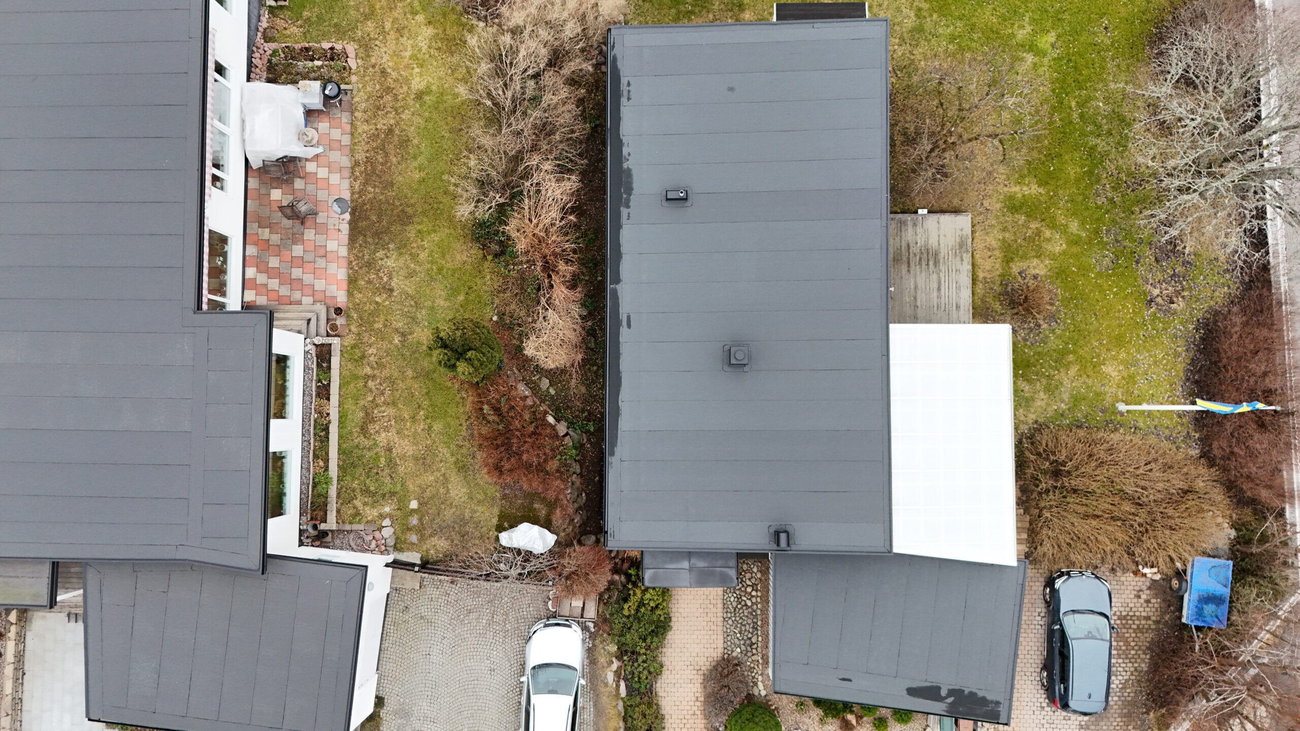 Fågelvy över ett platt hustak av asfaltpapp. Runt huset syns hustomten, två parkerade bilar samt ett närliggande grannhus.