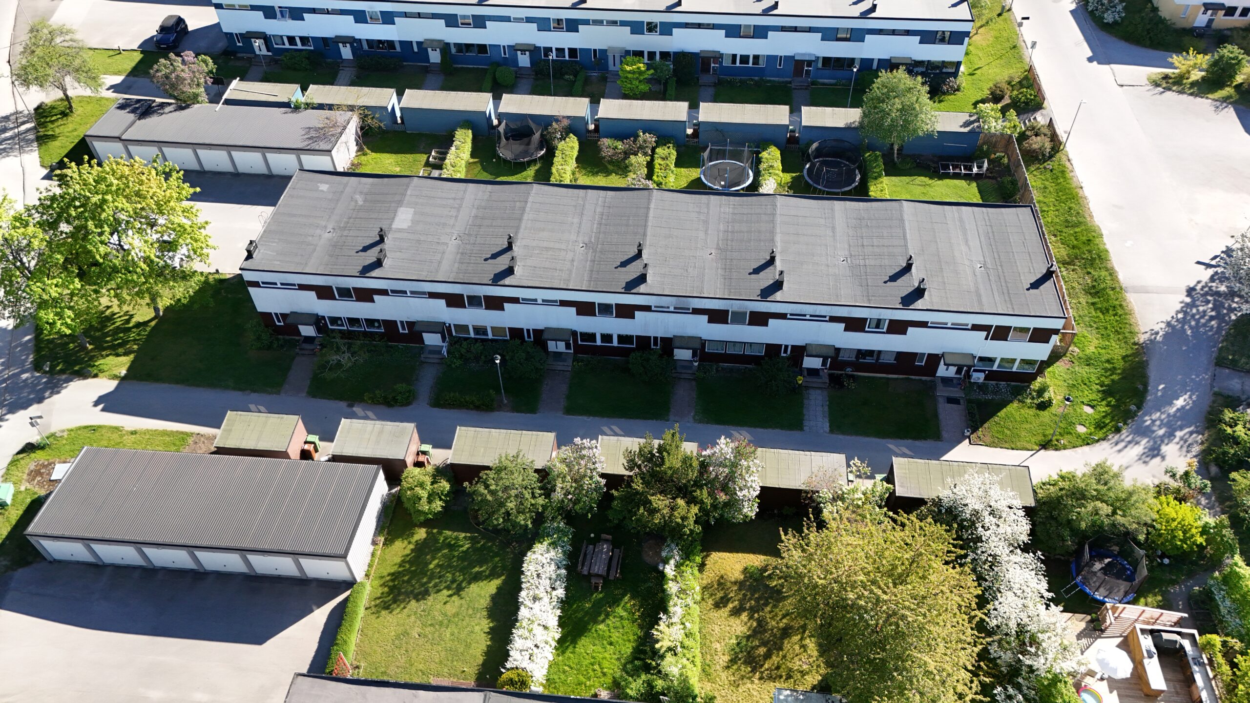 Fågelvy ovanifrån ett radhusområde. Radhusen har platt papptak. Runt husen finns det gräsmattor och träd, samt tillhörande garage.