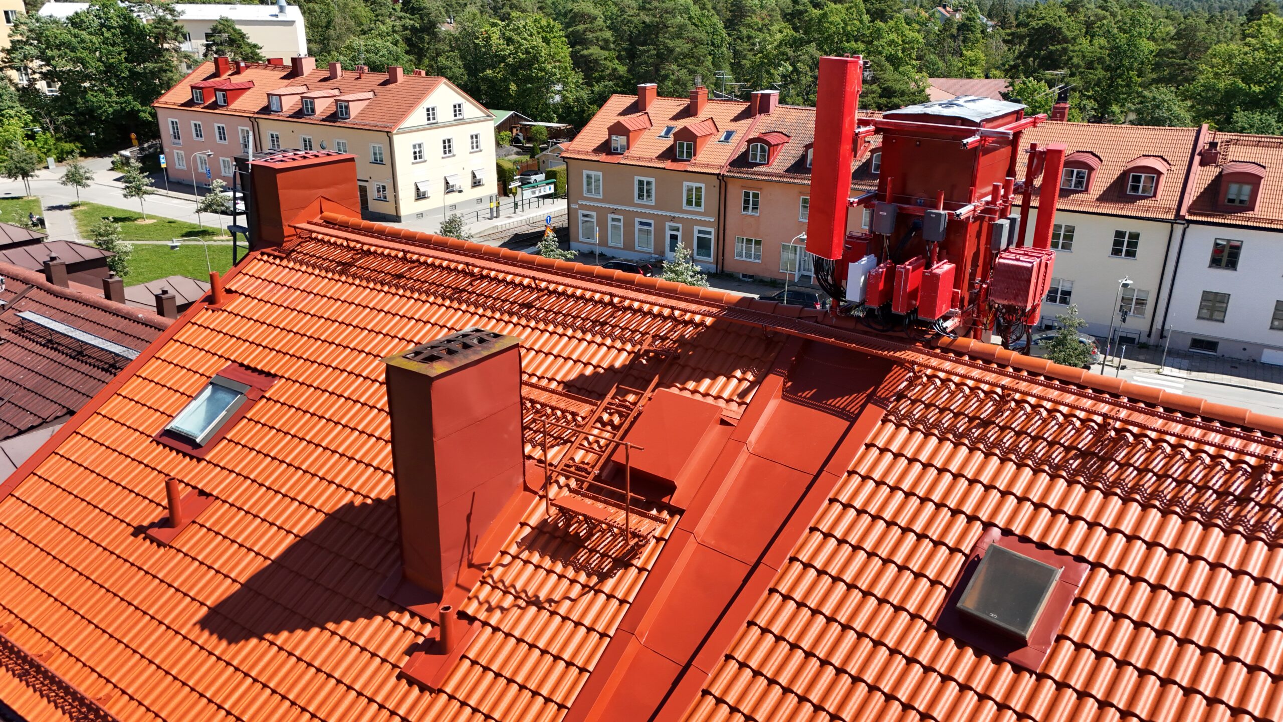 Närbild på hustaket på ett lägenhetshus. Taket är gjort av tegelpannor och plåt, med två skorstenar och två fönster. I bakgrunden syns anda lägenhetslängor med liknande tak.