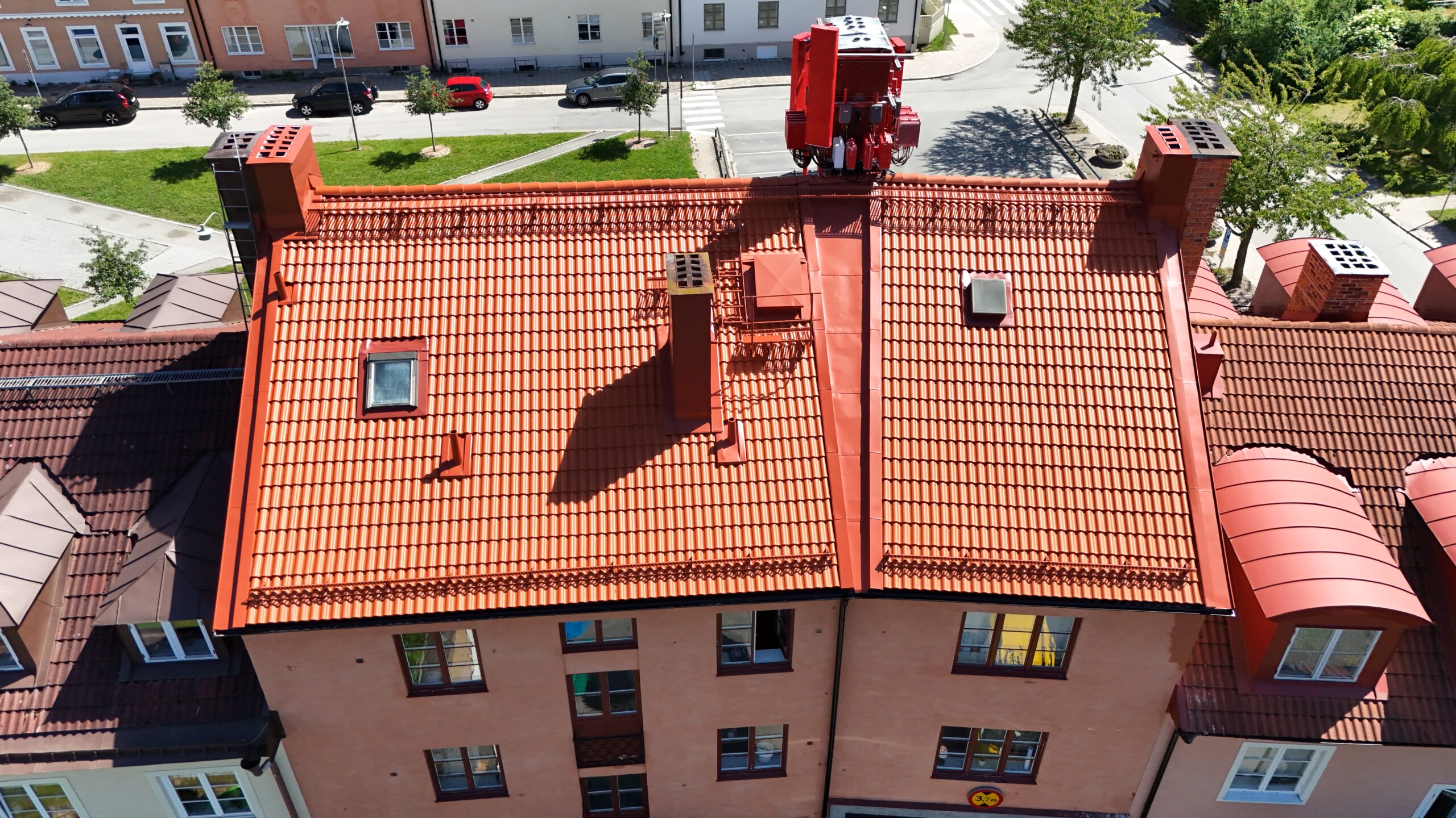 Fågelvy över ett lägenhetshus i ett husområde, med ett kombinerat tak av tegelpannor och plåt. På taket finns ett flertal skorstenar samt ett fönster. Husfasaden är putsad med ett flertal fönster.