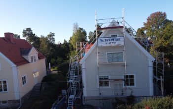 Fågelvy över hustak under takomläggning. Runt huset står flera byggställningar. I bakgrunden syns närliggande grannhus.