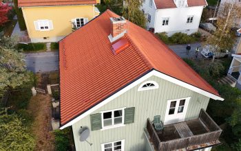 Fågelvy över en grön villa med sluttande tak av tegelpannor. På taket finns en skorsten i tegel samt snörasskydd. I bakgrunden syns närliggande grannhus.