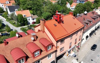 Fågelvy över en länga av lägenhetshus. Husens tak skiljer sig åt i färg och material. Bakom lägenhetshusen finns ett stort villaområde och stora träd.