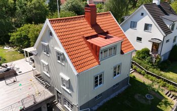 Fågelvy över en ljusgrå villa med kombinerat hustak. Taket består av tegeltak och bandtäckt plåt, med en skorsten i plåt. Villan har en stor altan runt sig.