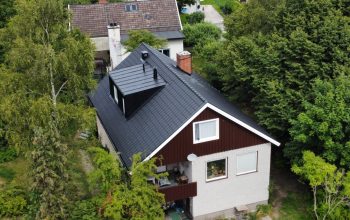 Fågelvy över ett stor hus i tegel och trä. Hustaket lutar och är gjort av svarta takpannor. Fågelvy över ett stor hus i tegel och trä. Hustaket lutar och är gjort av svarta takpannor. Intill huset står en röd bil parkerad.